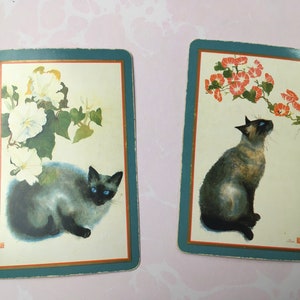 May include: Two vintage playing cards featuring Siamese cats. One card shows a cat with white flowers, the other with red flowers. Both cards have a teal border and a cream background.