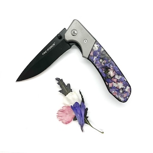 May include: A black folding knife with a silver handle and a blade marked "TAC-FORCE". The handle features a colorful floral pattern. A small arrangement of dried flowers in pink, white, and blue is next to the knife.
