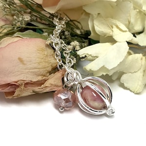May include: A silver chain necklace with a pink and white marble charm inside a silver cage. A pink crystal bead hangs from the cage.