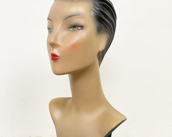 Könnte beinhalten: Eine Vintage-Büstenpuppe mit schwarzem Sockel. Die Puppe hat ein modelliertes Gesicht mit bemalten Gesichtszügen, darunter rote Lippen und blaue Augen. Die Puppe hat schwarzes Haar, das zu einer eleganten, modernen Hochsteckfrisur gestylt ist.