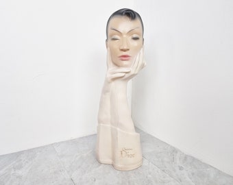 Könnte beinhalten: Ein weißer Gips-Mannequin-Kopf mit geschlossenen Augen und Händen, die auf dem Kinn ruhen. Der Mannequin trägt an jeder Hand einen weißen Handschuh. Die Basis des Mannequins ist mit dem Text "Christian Dior" beschriftet.