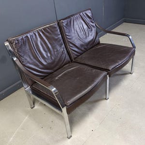 May include: A two-seater sofa with a dark brown leather seat and backrest. The frame is made of polished chrome. The sofa has a minimalist design with straight lines and a modern aesthetic. The sofa is suitable for a living room or office.