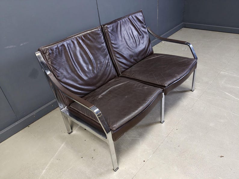 May include: A two-seater sofa with a dark brown leather seat and backrest. The frame is made of polished chrome. The sofa has a minimalist design with straight lines and a modern aesthetic. The sofa is suitable for a living room or office.