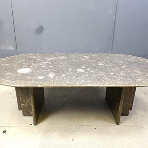 May include: A large, oval-shaped coffee table with a speckled gray stone top. The table has a dark brown base with a geometric design. The table is approximately 60 inches long and 30 inches wide.