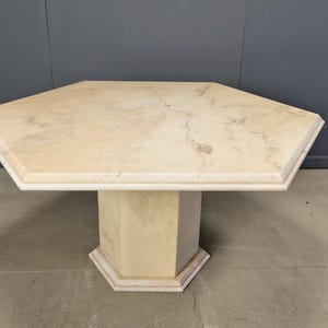Op de afbeelding: Een achthoekige tafel met een beige marmeren blad. De tafel heeft een bijpassende achthoekige voet. De tafel is ongeveer 122 cm breed en 76 cm hoog.
