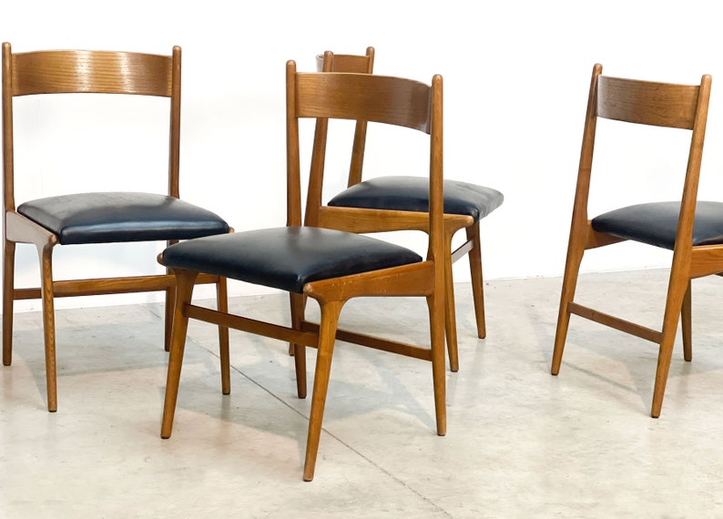 May include: Four wooden chairs with black leather seats. The chairs have a mid-century modern design, featuring a light brown wood frame and a simple, elegant silhouette. The chairs are arranged in a group, showcasing their design and style.
