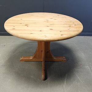 May include: A circular wooden table in a light brown hue. It features a central pedestal base with four legs. The tabletop is smooth and seems to be made of solid wood. The table is positioned against a grey and blue backdrop.