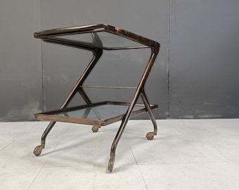 Op de afbeelding: Een tweelaagse metalen serveerwagen met glazen planken en wielen. De wagen is gemaakt van donker metaal met een geborstelde afwerking.