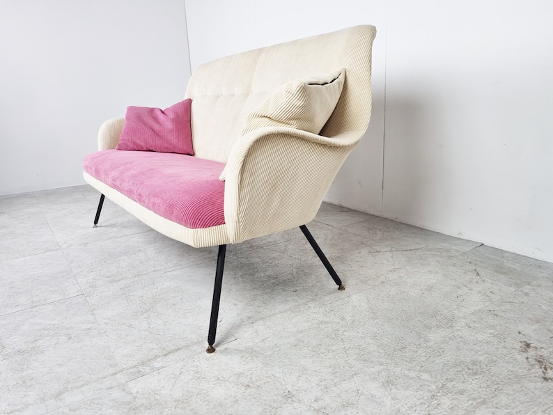 May include: A white and pink corduroy sofa with black metal legs. The sofa has two pillows, one pink and one white.
