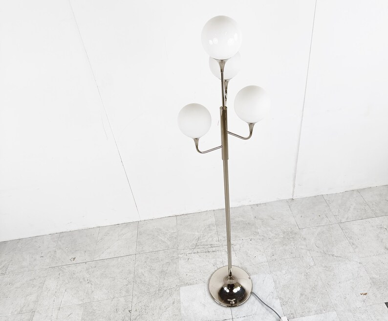 May include: A silver floor lamp with four white globe-shaped shades. The lamp has a long, slender stem and a circular base.