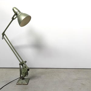 Könnte beinhalten: Eine architektenlampe aus salbeigrünem metall mit verstellbarem gelenkarm, quadratischem fuß und kuppelförmigem schirm. Die lampe verfügt über sichtbare mechanische gelenke und ein schwarzes stromkabel, das aus dem fuß herausführt.
