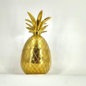 Op de afbeelding: Een metallic goudkleurig ananasvormig decoratieobject met een gepolijste afwerking. Het oppervlak is voorzien van een herhalend ruitvormig patroon over het lichaam en een reeks puntige, rechtopstaande bladeren aan de bovenkant van de structuur.