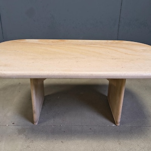 Pode incluir: Uma mesa de centro retangular de cor clara com cantos arredondados. A mesa tem um padrão de grão de madeira clara e é suportada por duas pernas angulares em estilo pedestal. A mesa tem cerca de 122 cm de comprimento.
