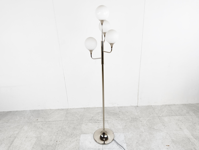 May include: A silver floor lamp with five white globe-shaped shades. The lamp has a long, slender stem and a round base.