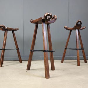 Può includere: Tre sgabelli da bar in legno con finitura scura. Ogni sgabello ha una seduta a forma di sella e un design scolpito unico nella parte superiore. Gli sgabelli hanno un design a gambe triangolari e un poggiapiedi in metallo. Gli sgabelli sono alti circa 76 cm.