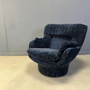 Puede incluir: Un sillón de felpa azul oscuro con base redondeada y respaldo alto. La silla tiene una textura suave y aterciopelada y presenta grandes reposabrazos curvos. El diseño general es moderno y cómodo, adecuado para una sala de estar o un área de descanso.