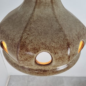 May include: A beige ceramic pendant light fixture with a textured surface and multiple cut-out holes. The light is on and illuminates the holes from the inside.