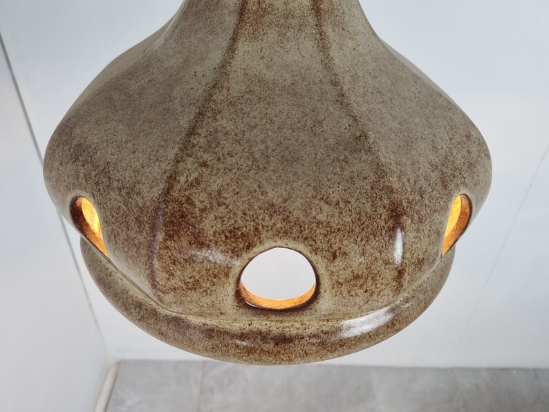 May include: A beige ceramic pendant light fixture with a textured surface and multiple cut-out holes. The light is on and illuminates the holes from the inside.