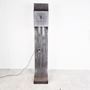 Könnte beinhalten: Eine hohe, silberfarbene Uhr mit geometrischem Design. Die Uhr hat ein rundes Zifferblatt mit schwarzen Zeigern und ein Pendel.