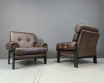May include: Two brown leather armchairs with black frames. One chair has a button-tufted back and rolled arms. The other chair has a high back with a cylindrical headrest and a cushioned seat. The chairs are placed against a gray wall.