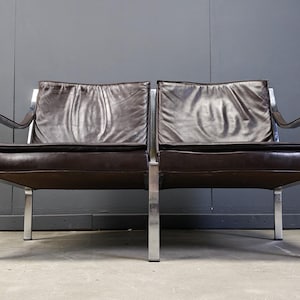 May include: A mid-century modern two-seater sofa with a dark brown leather seat and back cushions. The frame is made of polished chrome, with a minimalist design. The sofa is set against a gray wall.