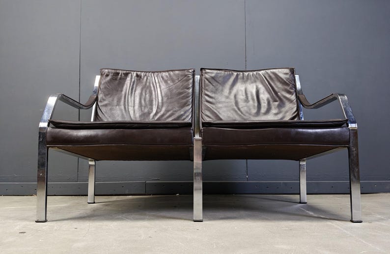 May include: A mid-century modern two-seater sofa with a dark brown leather seat and back cushions. The frame is made of polished chrome, with a minimalist design. The sofa is set against a gray wall.