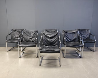 May include: Six black leather armchairs with chrome frames are arranged in a room. The chairs have a modern design, with a sleek, minimalist aesthetic. The leather appears to be slightly wrinkled.
