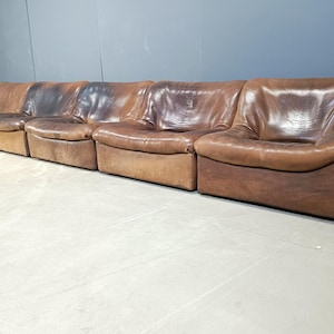 Op de afbeelding: Een bruine leren modulaire bank met vijf afzonderlijke zitplaatsen. De bank is gemaakt van distressed leer met een verweerde look.