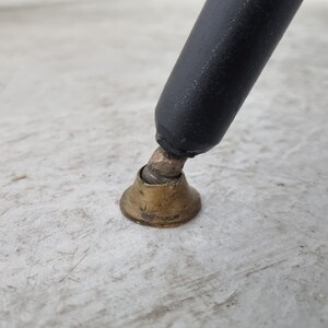 May include: Close-up of a black metal chair leg with a brass foot cap. The foot cap is round and has a slightly flared edge.