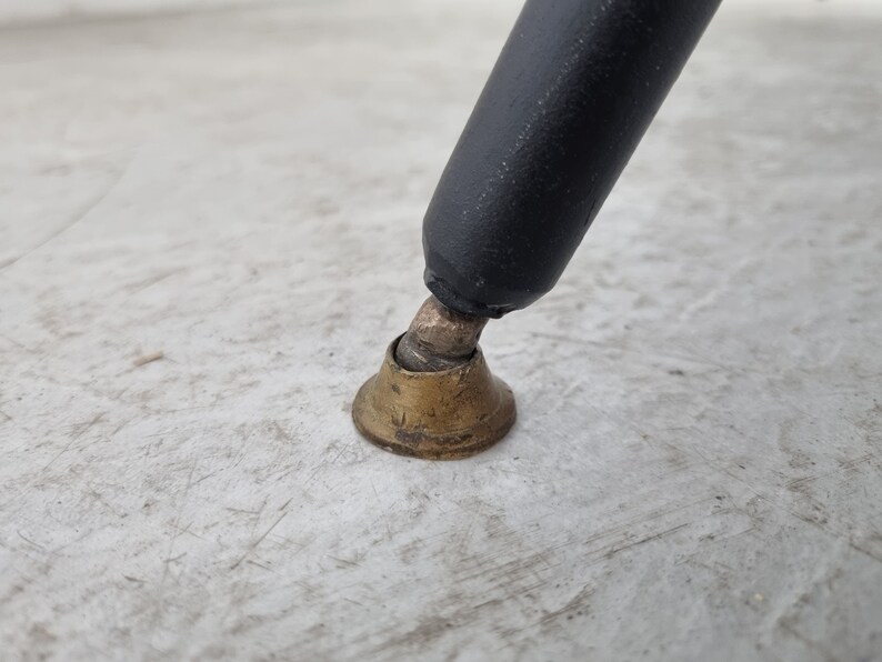 May include: Close-up of a black metal chair leg with a brass foot cap. The foot cap is round and has a slightly flared edge.
