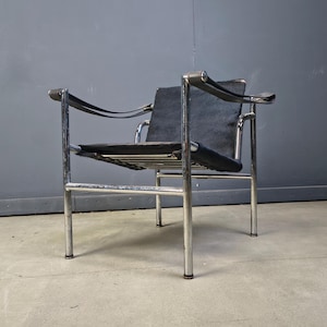 May include: A modern black leather and chrome armchair. The chair features a rectangular seat and backrest, with curved armrests and a tubular chrome frame. The chair is set against a gray wall and concrete floor.