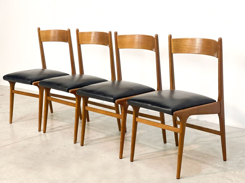 May include: Four mid-century modern dining chairs. The chairs have a light brown wooden frame with a curved backrest and black leather seats. The legs are tapered and angled, and the chairs are arranged in a row.