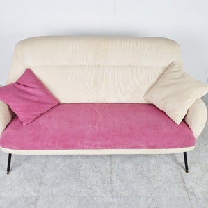 May include: A white and pink corduroy sofa with two pillows. The sofa has a curved back and black metal legs.