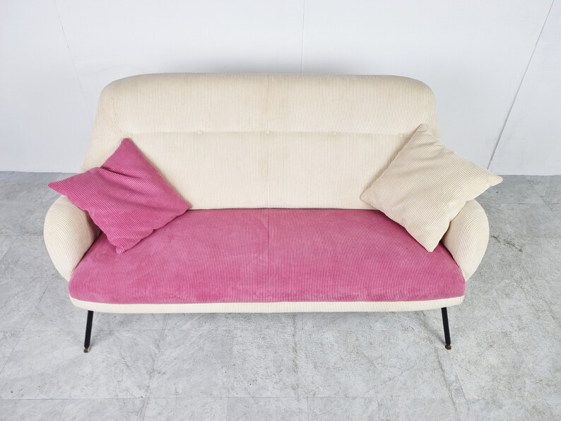 May include: A white and pink corduroy sofa with two pillows. The sofa has a curved back and black metal legs.