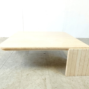 May include: A large, rectangular, travertine coffee table with a rounded edge and a unique, geometric base. The table is made of natural stone and has a light beige color.