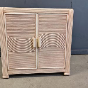 Puede incluir: Un mueble de dos puertas de color claro, hecho de ratán tejido. Las puertas tienen líneas horizontales con un diseño curvo en el centro. El mueble tiene forma rectangular, con una parte superior e inferior planas y dos pequeños tiradores.