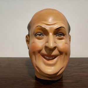 Puede incluir: Un busto de yeso vintage de un hombre calvo, sonriendo con la boca abierta, mostrando los dientes. El busto está pintado en tonos marrones y beige.