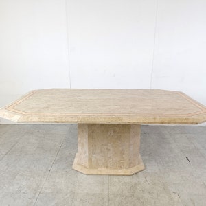 Puede incluir: Una mesa de comedor rectangular grande de travertino con un borde biselado y una base de pedestal único. La superficie de la mesa tiene un color beige claro con un patrón sutil de vetas marrones y blancas.