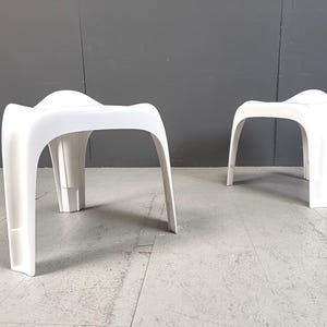 May include: Two white plastic stools with a unique, curved design. The stools are made of a smooth, white plastic and have a simple, modern aesthetic.