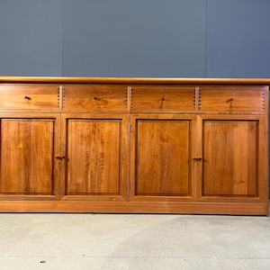 May include: A large, wooden sideboard with a warm, reddish-brown finish. It features four cabinet doors and four drawers with small, round knobs. The top has a slightly rounded edge. The piece is set against a dark blue wall.
