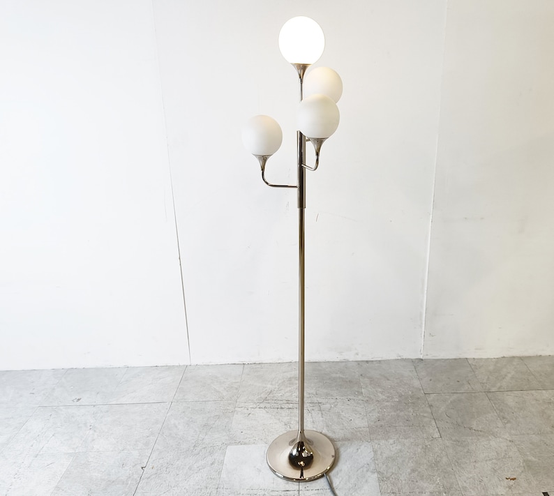 May include: A chrome floor lamp with five white globe-shaped shades. The lamp has a tall, slender base and a curved arm that extends to the side.