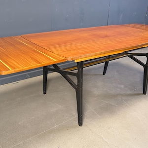 Peut inclure: Une table à manger rectangulaire dotée d'un plateau en bois poli aux tons chauds avec une rallonge. La table est soutenue par une structure noire mate foncée composée de quatre pieds fuselés et de traverses de renfort interconnectées.