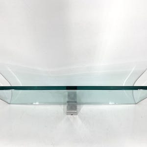 May include: A clear glass shelf with a rectangular shape and beveled edges. The shelf is supported by a silver-colored metal bracket. The shelf is designed to be mounted on a wall.