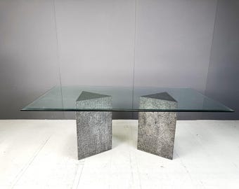 May include: A glass-top dining table with two gray stone bases. The table is rectangular and has a clear glass top. The bases are made of a textured stone and are triangular in shape.