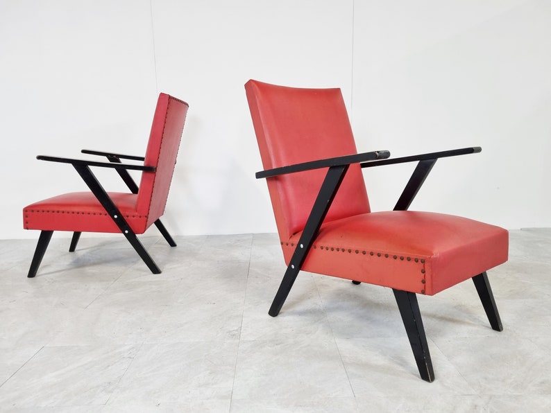 May include: Two red upholstered armchairs with black wooden frames. The chairs have a mid-century modern design with angled legs and arms.