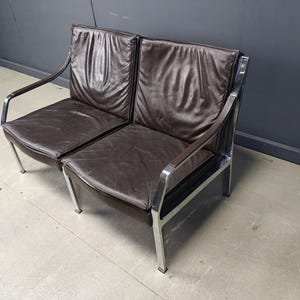 May include: A two-seater brown leather sofa with a chrome frame. The cushions are a dark brown, and the frame has a curved armrest design. The sofa has a mid-century modern aesthetic.