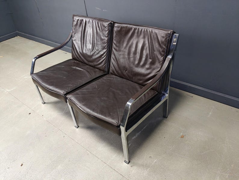 May include: A two-seater brown leather sofa with a chrome frame. The cushions are a dark brown, and the frame has a curved armrest design. The sofa has a mid-century modern aesthetic.