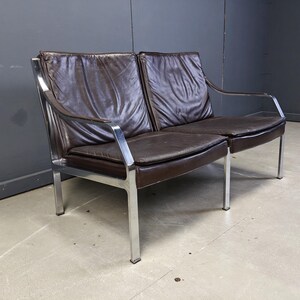 May include: A mid-century modern two-seater sofa with a dark brown leather seat and back. The frame is made of polished chrome, with a minimalist design. The sofa has a low profile and a sleek, contemporary look.