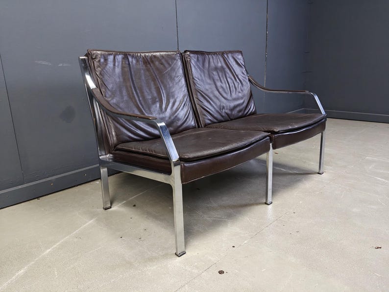 May include: A mid-century modern two-seater sofa with a dark brown leather seat and back. The frame is made of polished chrome, with a minimalist design. The sofa has a low profile and a sleek, contemporary look.