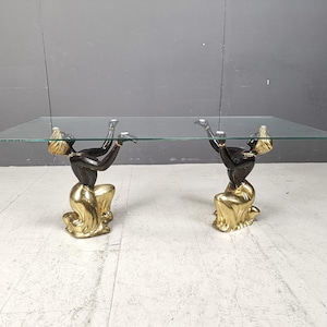 May include: A glass-topped coffee table with a distinctive design. The base consists of two figures, with black bodies and gold-coloured clothing, kneeling and supporting the glass top. A unique piece of furniture, the table is approximately 150 cm long.